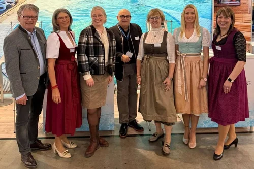 Group at Poolside Event A group of seven individuals dressed in traditional attire standing beside a swimming pool backdrop. They are smiling, appearing at an event or exhibition, with various hairstyles and accessories.