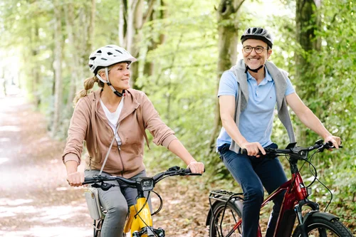 Zwei Personen fahren mit E-Bikes auf einem Waldweg. Sie tragen Helme und lächeln, während sie miteinander sprechen. Bäume und grünes Laub umgeben den Weg.