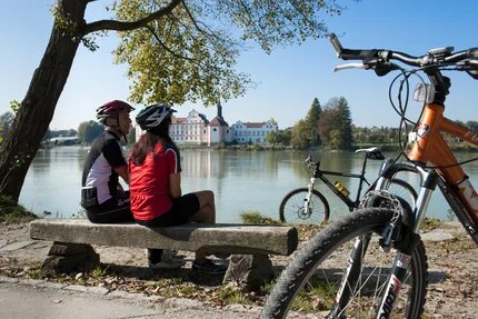 Ein Radfahrer-Paar sitzt auf einer Bank am Flussufer und blickt auf ein Schloss im Hintergrund. Fahrräder stehen im Vordergrund neben ihnen.