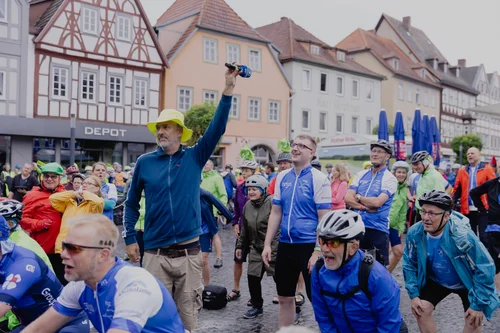 Radveranstaltung mit jubelndem Publikum Ein Mann in einem blauen Pullover und einem gelben Hut hält ein Mikrofon hoch, umgeben von einer Gruppe von Radfahrern in bunten Trikots. Im Hintergrund sind historische Gebäude zu sehen. Viele Zuschauer lächeln und schauen auf ihn.