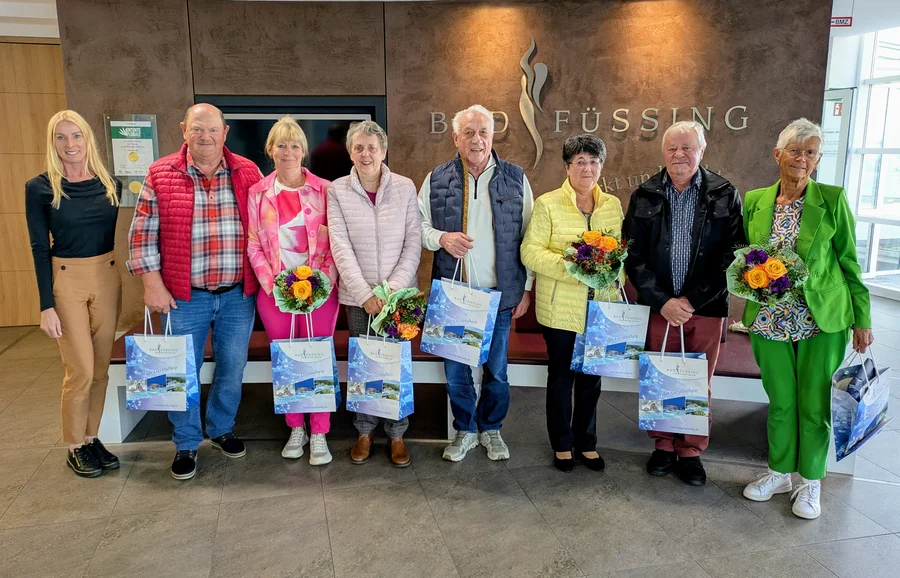Gruppe in der Bad-Füssing-Lobby Sieben ältere Personen und eine jüngere Frau stehen in einer Lobby vor der Beschriftung "Bad Füssing" und halten Geschenktüten sowie Blumensträuße in den Händen.