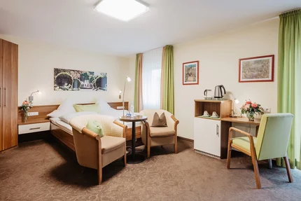 Gemütliches Hotelzimmer Ein gemütliches Hotelzimmer mit einem Doppelbett, zwei Sesseln, einem Tisch und einer kleinen Kaffee-/Teeküche. An der Wand hängen zwei Bilder. Vor dem Fenster stehen Vorhänge in Gelb und Grün.