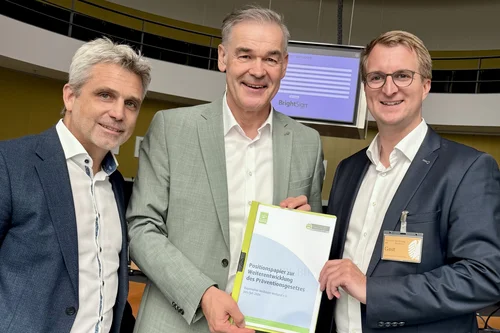 Drei Männer mit Positionspapier Drei Männer stehen in einem Raum und halten ein Positionspapier. Jeder Mann trägt Anzugskleidung und hat ein freundliches Lächeln. Im Hintergrund ist ein Bildschirm sichtbar.