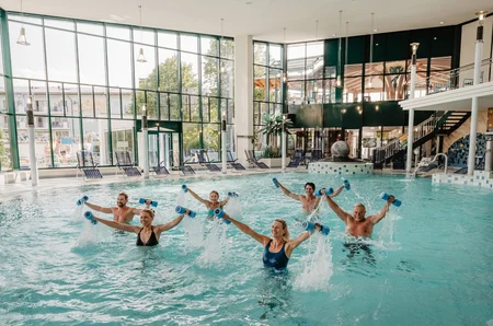 Eine Gruppe von sechs Personen macht Aquagymnastik im Wasser, während sie kleine blaue Gewichte halten. Der Pool ist hell erleuchtet mit großen Fenstern im Hintergrund.