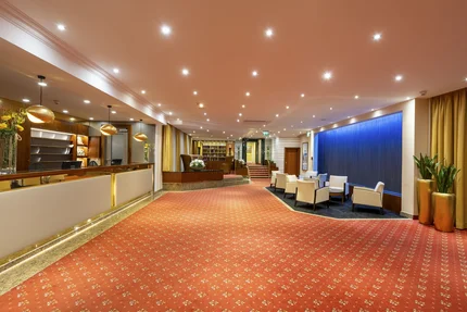 Eine elegante Hotellobby mit einem Empfangstresen, Sitzbereichen und modernen Einrichtungen. Zur linken Seite sind Pflanzen und dekorative Lichtquellen sichtbar. Der Boden ist mit rot gemustertem Teppich ausgelegt.