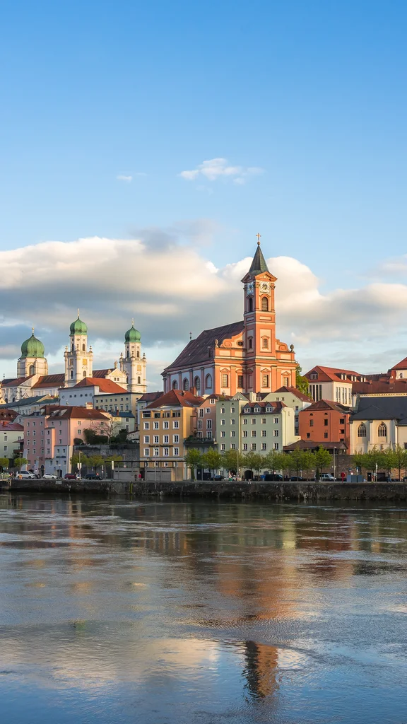 Flussstadt mit Kirchenansicht Eine Stadtansicht auf der linken Seite des Gerinnes mit bunten Gebäuden und Kirchen mit grünen Kuppeln im Hintergrund. Der Himmel zeigt einige Wolken und das Licht ist sanft.