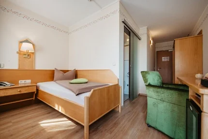 Ein einfaches Hotelzimmer mit einem Bett, einem Schreibtisch und einem Stuhl. An der Wand befindet sich eine Lampe. Eine Glastür führt zu einem Badezimmer.