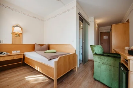 Ein einfaches Hotelzimmer mit einem Bett, einem Schreibtisch und einem Stuhl. An der Wand befindet sich eine Lampe. Eine Glastür führt zu einem Badezimmer.