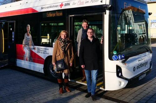 Personen vor einem Reisebus Drei Personen stehen vor einem Bus mit Werbung für Seniorenreisen. Der Bus hat eine offene Tür, und eine Frau mit einem Schildratschatten ist im Hintergrund sichtbar.