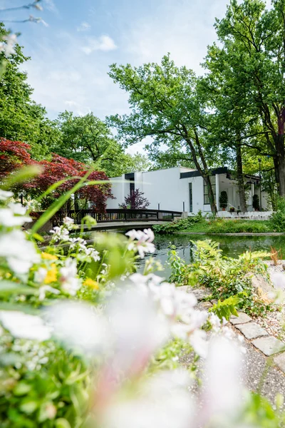 Ein ruhiger Garten mit einem Teich, umgeben von Bäumen und bunten Blumen. Im Hintergrund sieht man ein modernes Gebäude mit einem Bereich zum Sitzen.