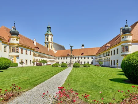 Historischer Innenhof mit Statue Ein großer Innenhof eines historischen Gebäudes, umgeben von gepflegten Rasenflächen und Büschen. In der Mitte steht eine Statue, flankiert von architektonischen Elementen und einem Turm im Hintergrund.