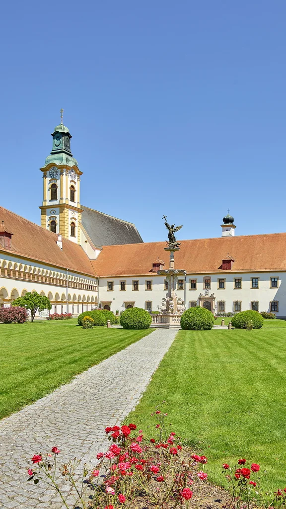 Ein großer Innenhof eines historischen Gebäudes, umgeben von gepflegten Rasenflächen und Büschen. In der Mitte steht eine Statue, flankiert von architektonischen Elementen und einem Turm im Hintergrund.