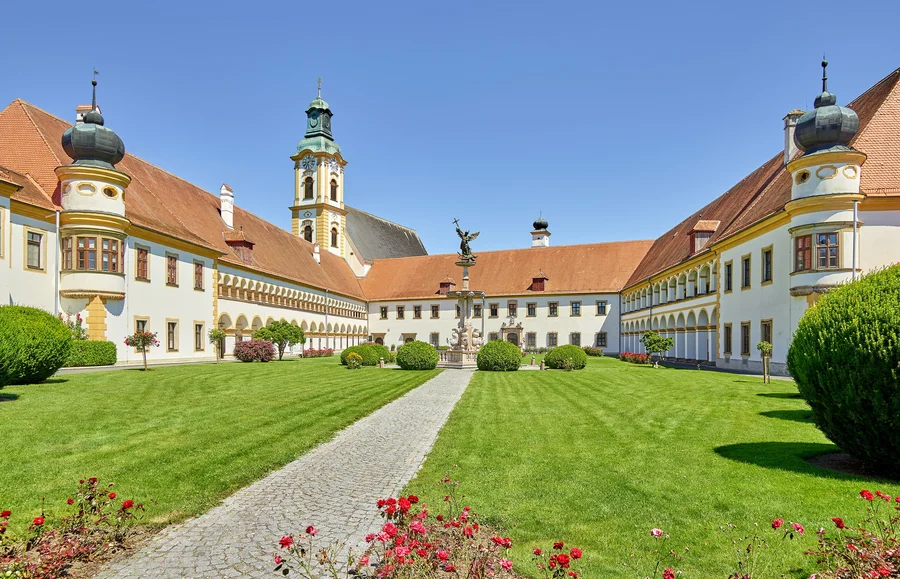 Ein großer Innenhof eines historischen Gebäudes, umgeben von gepflegten Rasenflächen und Büschen. In der Mitte steht eine Statue, flankiert von architektonischen Elementen und einem Turm im Hintergrund.