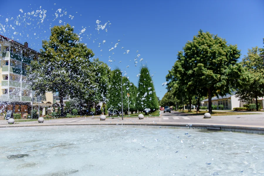 Spritzender Brunnen im Park Ein Brunnen mit spritzendem Wasser, umgeben von Bäumen und Gebäuden, einer klaren blauen Himmel im Hintergrund.