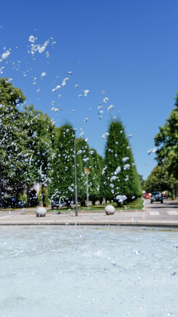 Ein Brunnen mit spritzendem Wasser, umgeben von Bäumen und Gebäuden, einer klaren blauen Himmel im Hintergrund.