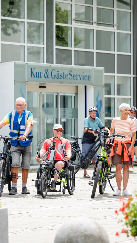 Gruppe von Radfahrern vor Kur-Service Eine Gruppe von radfahrenden Menschen steht vor dem Kur- und GästeService. Sie haben verschiedene Fahrräder, einschließlich eines speziellen Fahrrads für Menschen mit Behinderung. Blumen und moderne Architektur sind im Hintergrund zu sehen.
