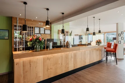 Moderne Empfangstheke im Empfangsbereich Eine moderne Empfangstheke mit Holzverkleidung, Pflanzenarrangements und hängenden Lampen in einem hellen Raum. Auf einem Tisch stehen Informationsmaterialien und Getränke.