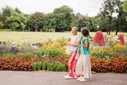 Zwei Frauen gehen lächelnd in einem Park. Bunte Blumen umgeben den Gehweg, während im Hintergrund andere Parkbesucher auf Bänken sitzen.