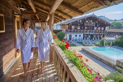 Zwei Frauen in Bademänteln laufen auf einer Holzveranda und lächeln. Im Hintergrund sieht man einen Pool und blühende Blumen.