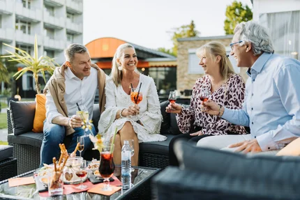 Eine Gruppe von vier Personen sitzt an einem Tisch mit Snacks und Getränken. Sie unterhalten sich fröhlich auf einer Terrasse mit Pflanzen im Hintergrund.
