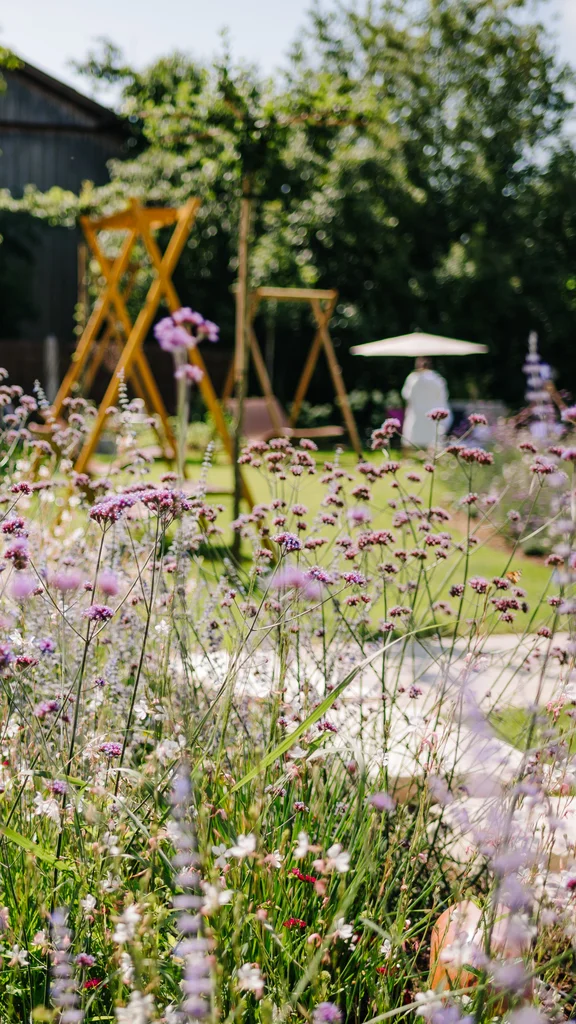 Ein sonniger Garten mit bunten Blumen, Schaukeln und Bäumen. Im Hintergrund sieht man einige Menschen und eine Terrasse.