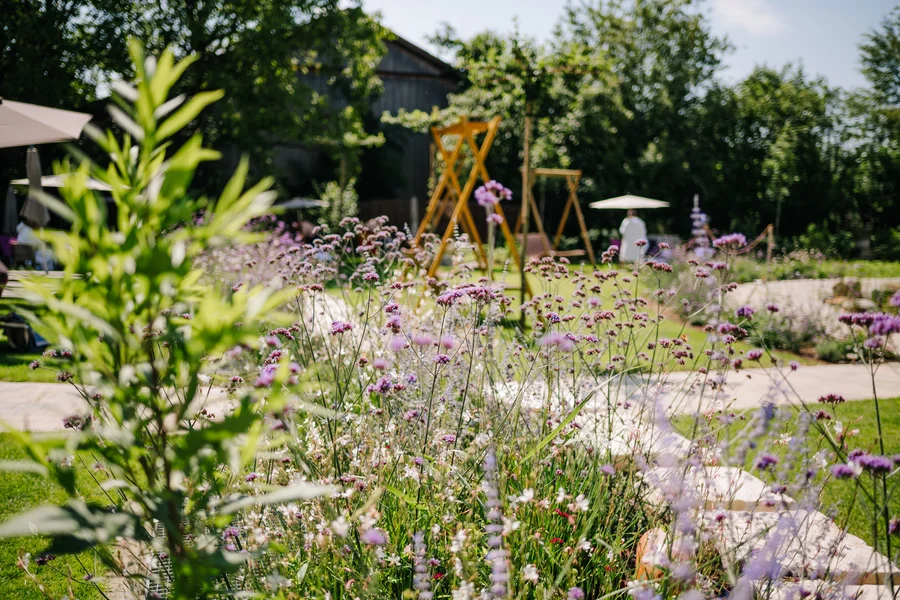 Ein sonniger Garten mit bunten Blumen, Schaukeln und Bäumen. Im Hintergrund sieht man einige Menschen und eine Terrasse.