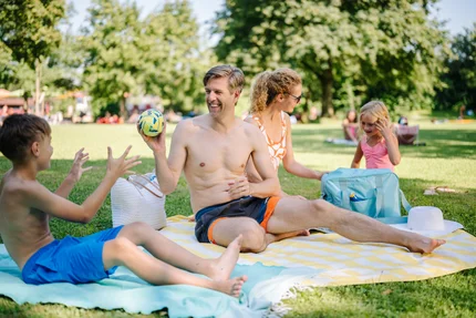 Eine Familie hat ein Picknick im Park. Ein Mann sitzt auf einer Decke, hält einen Ball und spielt mit einem Jungen. Eine Frau und ein kleines Mädchen sind nebendran beschäftigt.