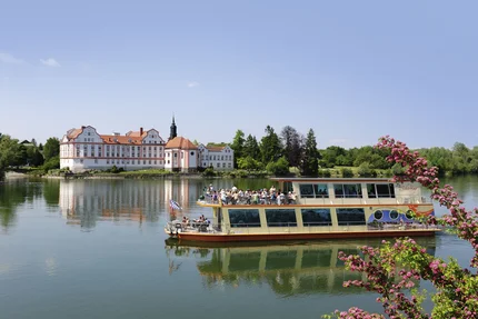 Eine malerische Landschaft mit einem Schloss im Hintergrund und einem Ausflugsboot auf dem Wasser. Blühende Pflanzen rahmen die Szene ein.