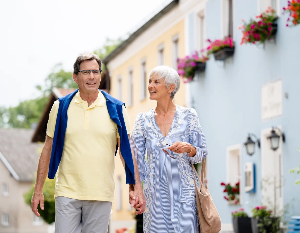 Älteres Paar in buntem Dorf Ein älteres Paar läuft Hand in Hand in einer farbenfrohen Straße mit Blumen an den Fenstern. Der Mann trägt ein gelbes Shirt und eine blaue Jacke, die Frau ein gestreiftes Kleid mit Blumenmustern.