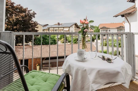 Balkon mit Tisch und Blumen Ein Balkon mit Tisch und Stühlen, dekoriert mit einer Vase und Blumen. Im Hintergrund sind Gebäude und Bäume sichtbar.