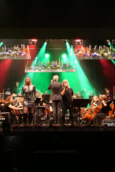 A live music performance featuring vocalists and an orchestra on stage, with colorful lights and floral decorations. The audience is visible in the foreground.