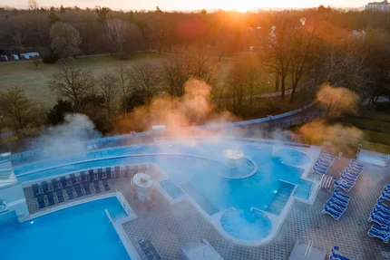 Außenpool bei Sonnenaufgang Ein Außenpoolbereich mit mehreren Becken, Liegen und aufsteigendem Dampf bei Sonnenaufgang. Im Hintergrund liegen Wiesen, Bäume und ein Park.