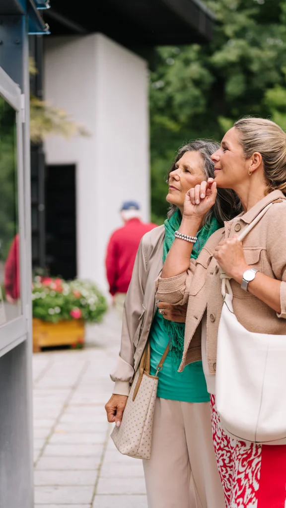 Frauen schauen sich Informationen an Zwei Frauen betrachten Schautafeln an einem Informationsschalter, umgeben von Blumen und Passanten. Eine Frau hat eine graue Jacke und eine grüne Bluse, die andere trägt eine beige Jacke und hält eine Tasche.