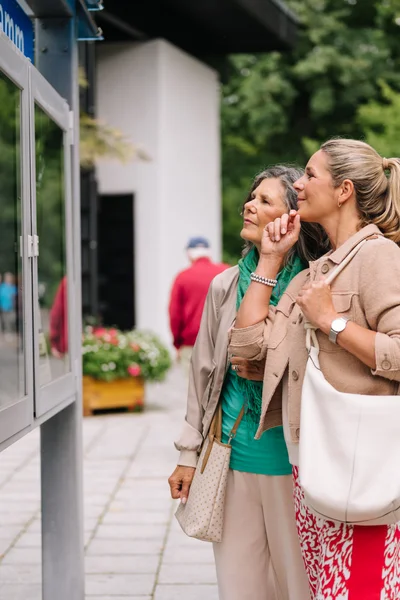 Frauen schauen sich Informationen an Zwei Frauen betrachten Schautafeln an einem Informationsschalter, umgeben von Blumen und Passanten. Eine Frau hat eine graue Jacke und eine grüne Bluse, die andere trägt eine beige Jacke und hält eine Tasche.