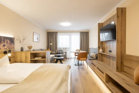 Ein modernes Hotelzimmer mit einem Bett und einem Wohnbereich. Es gibt einen Tisch mit Stühlen, eine graue Couch und ein Holzregal mit einem Fernseher. Natürliches Licht fällt durch die Vorhänge.