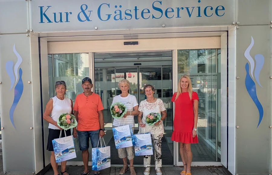 Fünf Personen stehen vor dem Kur- und GästeService, zwei Frauen halten Blumenstrauß, alle lächeln freundlich.