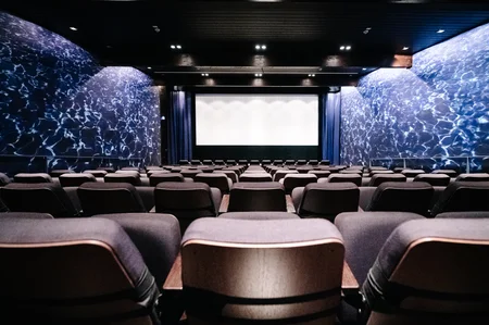 Leerer Kinosaal mit Leinwand Ein leerer Kinosaal mit gepolsterten Sitzen und einer großen Leinwand vorne. Die Wände sind mit einem blauen Wasserdesign verziert und die Beleuchtung erzeugt eine entspannte Atmosphäre.