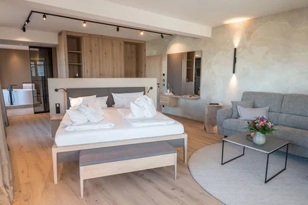 Ein modernes Hotelzimmer mit einem großen Doppelbett, einer Couch und einem kleinen Tisch. Ein Bad ist im Hintergrund sichtbar, zusammen mit Regalen. Helle Farben und natürliche Materialien dominieren den Raum.