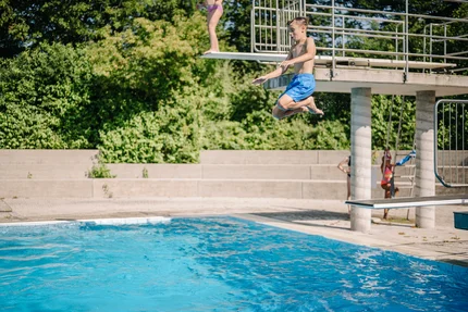 Ein Junge springt vom Sprungbrett in ein Schwimmbecken, während eine Person auf dem Sprungbrett steht. Im Hintergrund sind weitere Schwimmer und grüne Bäume sichtbar.