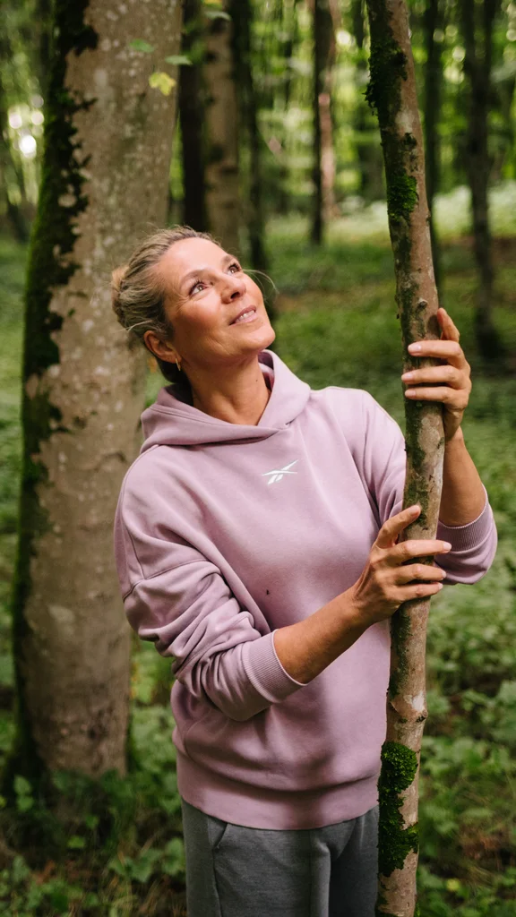 Frauen im Wald interagieren mit Bäumen Eine Frau steht im Wald und umarmt einen Baum. Im Hintergrund sind zwei weitere Frauen zu sehen, die ebenfalls eine Baumstütze berühren. Der Wald ist grün und reich an Bäumen.