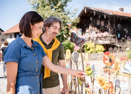 Besuch eines Kunsthandwerksmarkts Ein Mann und eine Frau stehen vor bunten Statuen und betrachten einen grünen Frosch. Der Mann trägt einen gelben Pullover um den Hals, die Frau hat ein blaues Kleid. Im Hintergrund ist ein traditionelles Holzhaus sichtbar. Menschen sind ebenfalls im Hintergrund.
