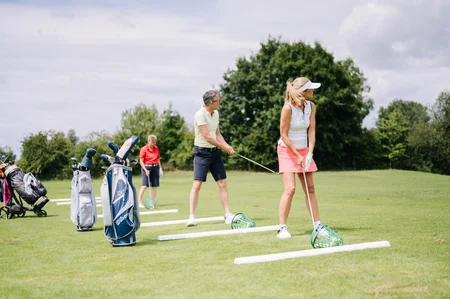 Golfspieler auf der Driving Range Drei Golfspieler auf einer Driving Range, zwei mit Schlägern in der Hand, ein dritter beobachtet. Golftaschen stehen in der Nähe.