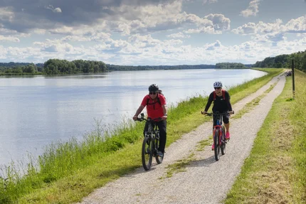 Ein Mann und eine Frau fahren Fahrrad entlang eines Flusses. Die Umgebung ist grün mit Bäumen, und der Himmel ist bewölkt. Es gibt einen Weg neben dem Wasser.