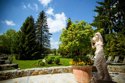 Ein Garten mit hohen Bäumen, einer Statue einer Frau, die einen Krug hält, und bunten Blumen in einem großen Topf.